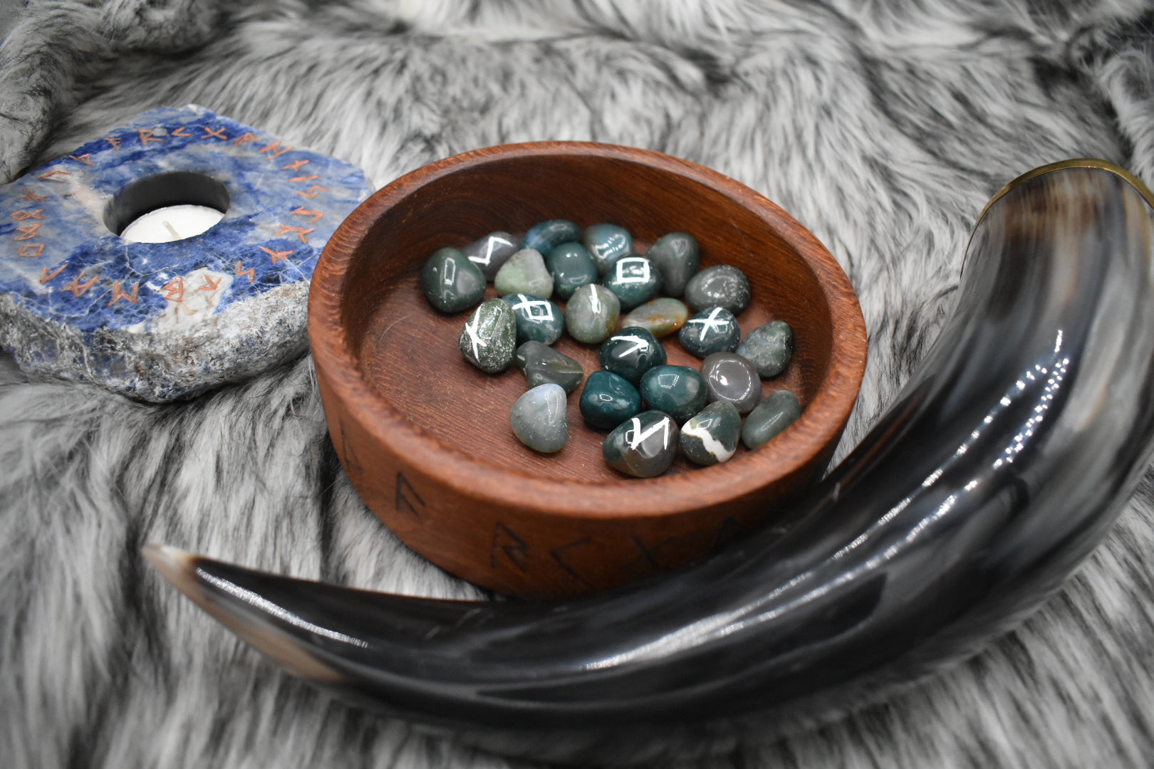 decorative photo of bloodstone elder futhark runes with a white inlay in a bowl on an altar with Viking drinking horn and sodalite candle holder